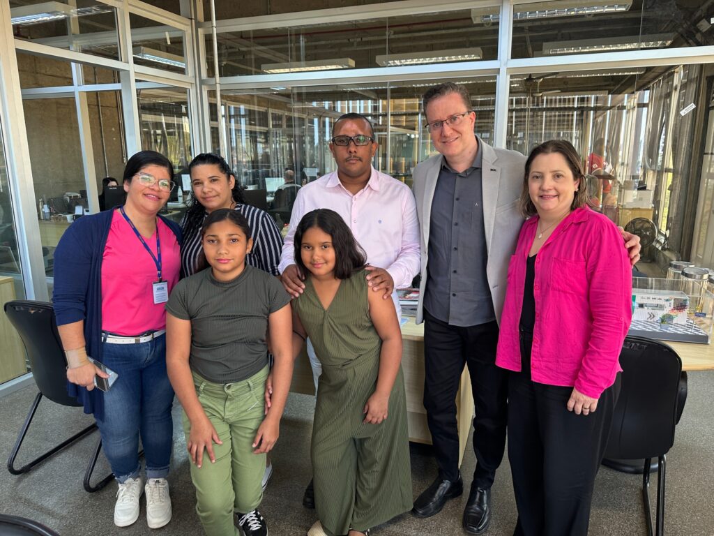 Um grupo de seis pessoas está posando para uma foto em um escritório. Há três adultos e duas crianças na frente. A primeira mulher da esquerda para a direita está usando um cardigã azul-marinho sobre uma blusa rosa choque. Ela tem óculos e está usando um crachá. Ela está usando jeans azul e tênis brancos. Ao lado dela está uma mulher com uma blusa listrada em preto e branco. Na frente delas, está uma menina com uma blusa verde justa e calças verdes. Ao lado dela, uma menina com um macacão verde. Atrás delas, um homem de camisa rosa com óculos e um homem de camisa cinza e blazer cinza. A mulher mais à direita está usando uma blusa rosa choque sobre uma blusa preta e calças pretas. Ela está usando um colar com um pingente.