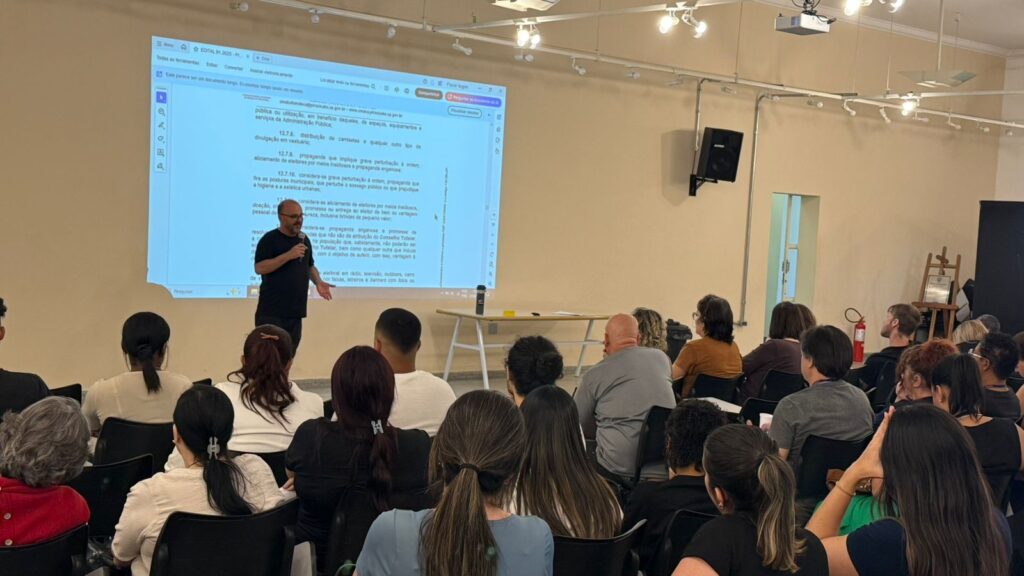 Um homem em pé, de camiseta preta, segura um microfone e fala diante de uma plateia em auditório amplo e iluminado, com teto moderno e várias luminárias acesas. À sua frente, uma tela projetada mostra o título “Responsabilidades da Equipe de Apoio” e uma lista de orientações sobre a organização das filas e verificação de documentos durante a eleição. O público, sentado em cadeiras escolares, acompanha atentamente.