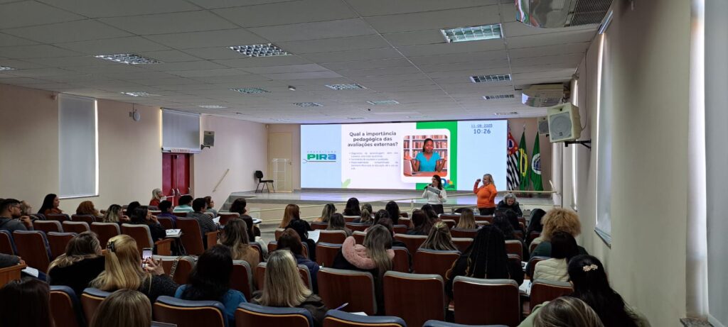A imagem mostra uma palestra em andamento em uma sala ampla, com diversas pessoas sentadas em cadeiras organizadas em fileiras. No telão à frente, é projetado um slide com o título “Qual a importância pedagógica das avaliações externas?”, seguido de três tópicos que destacam o papel dessas avaliações no diagnóstico da rede, no apoio à gestão escolar e na análise das turmas pelos professores. A apresentação ocorre no dia 11 de agosto de 2022, às 10h26, conforme indicado no slide. Dois palestrantes estão posicionados diante do público — um com camisa laranja e outro com camisa branca — enquanto bandeiras do Brasil e do estado de São Paulo decoram o lado direito do ambiente. A plateia parece atenta, com alguns participantes fazendo anotações ou utilizando dispositivos eletrônicos.