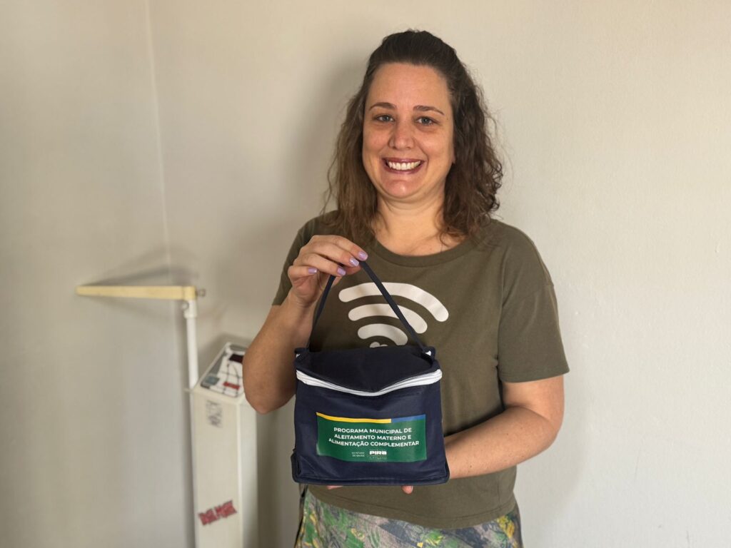 A mulher sorri para a câmera enquanto segura uma bolsa azul escura com alças. A bolsa tem um zíper branco e uma etiqueta retangular verde com texto amarelo e branco. O texto na etiqueta diz "PROGRAMA MUNICIPAL DE ALEITAMENTO MATERNO E ALIMENTAÇÃO COMPLEMENTAR" e também inclui um pequeno logotipo. A mulher está usando uma camiseta verde oliva com um grande símbolo branco de Wi-Fi no centro. O fundo é uma parede de cor clara com alguns equipamentos médicos à esquerda.