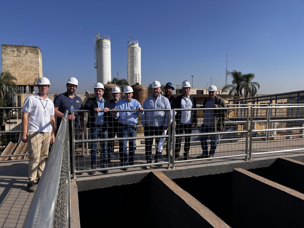 Oito homens, todos usando capacetes brancos, estão em pé em uma passarela metálica com grades. Eles estão em frente a duas grandes torres cilíndricas brancas com a palavra "SEMAE" escrita em letras pretas. Há um céu azul claro acima deles. O homem mais à esquerda está vestindo uma camisa polo branca e calças cáqui. Os outros homens estão vestindo camisas de manga comprida e calças jeans. O homem mais à direita está vestindo uma camisa xadrez e calças cáqui. Atrás deles, há um edifício de concreto e algumas árvores de palma.