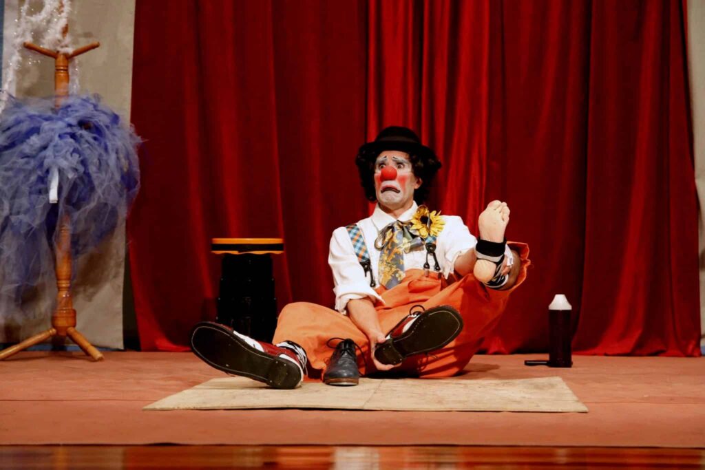 O palhaço Picuinha está sentado no chão do palco, em frente à cortina vermelha. Ele olha para frente com expressão triste e exagerada. Usa sapatos grandes pretos e brancos, calça laranja e camisa branca com adereços coloridos.
