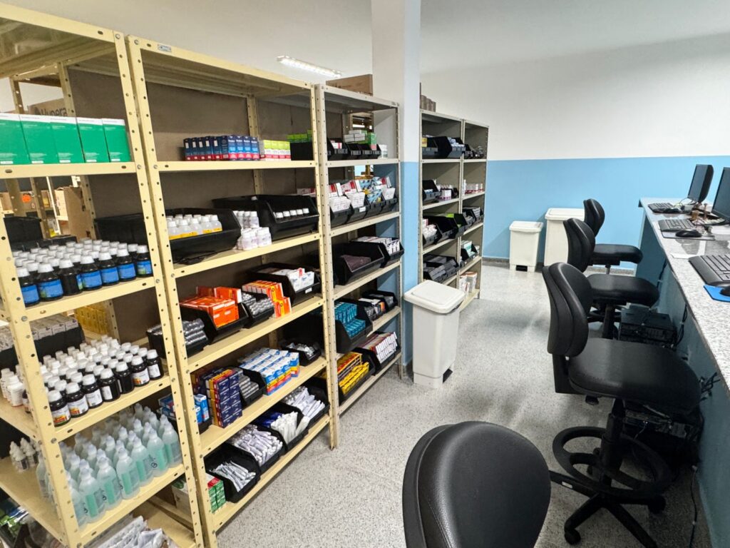  imagem mostra uma farmácia, com prateleiras cheias de caixas e frascos de medicamentos e suprimentos médicos. Há também uma área de trabalho com computadores e cadeiras, sugerindo que este é um local onde os medicamentos são dispensados ou gerenciados. As prateleiras estão organizadas com diferentes tipos de produtos, alguns em caixas coloridas e outros em frascos. Há também lixeiras em alguns pontos do ambiente.
