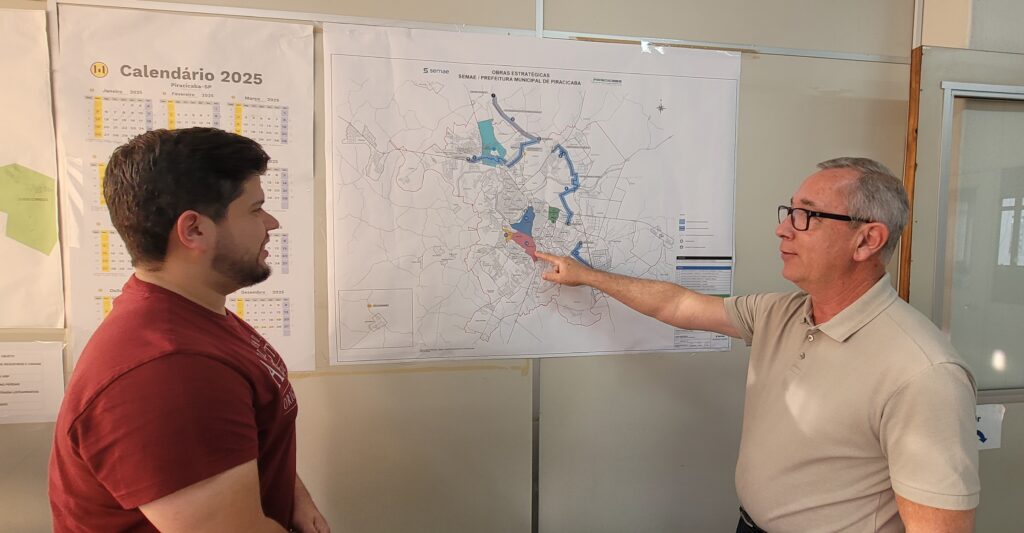Dois homens estão em pé diante de uma parede com vários mapas e gráficos. À esquerda, um homem mais jovem, de cabelo escuro e barba, veste uma camiseta vinho. Ele olha para a direita, na direção do outro homem. À direita, um homem mais velho, com cabelo grisalho curto e óculos, usa uma camisa polo bege clara. Ele aponta com o dedo indicador direito para um grande mapa na parede. O mapa tem o título "OBRAS ESTRATÉGICAS SEMAE / PREFEITURA MUNICIPAL DE PIRACICABA" e parece ser um plano estratégico de desenvolvimento urbano da cidade de Piracicaba. À esquerda do mapa, há um calendário de 2025 mostrando os meses de janeiro, fevereiro e março. Outros papéis ou gráficos estão na parede também, mas estão desfocados ou parcialmente cobertos.