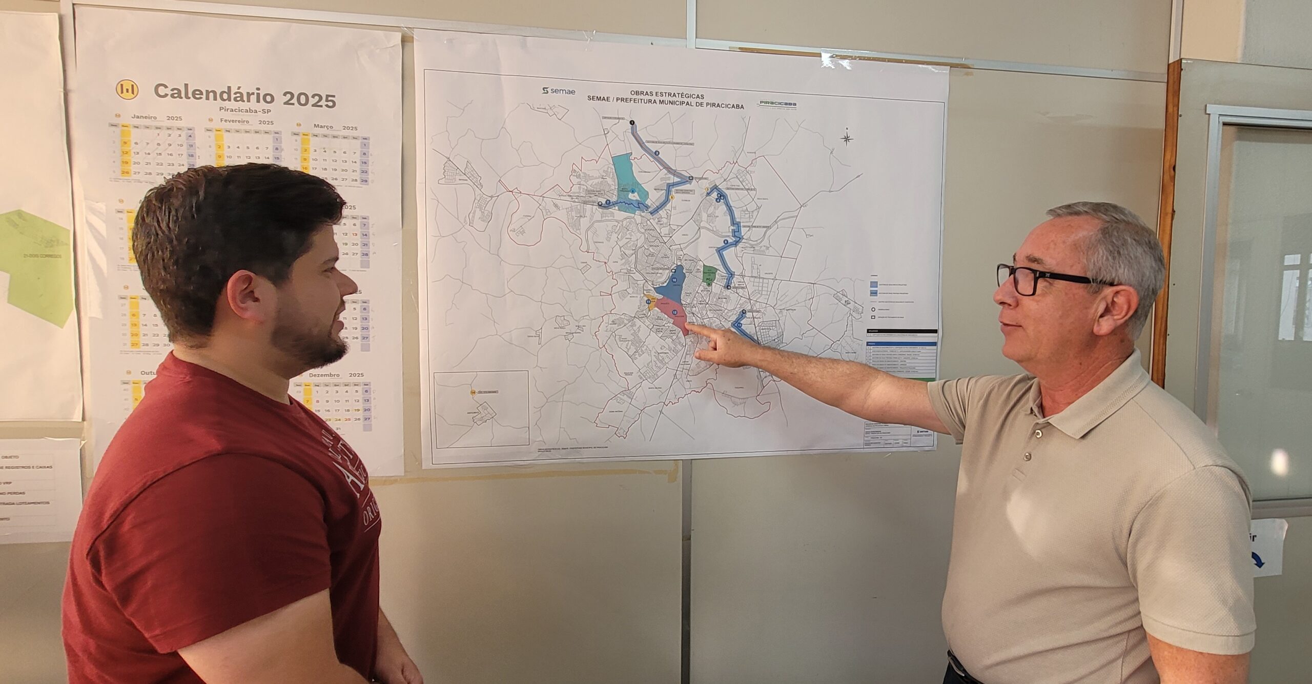 Dois homens estão em pé diante de uma parede com vários mapas e gráficos. À esquerda, um homem mais jovem, de cabelo escuro e barba, veste uma camiseta vinho. Ele olha para a direita, na direção do outro homem. À direita, um homem mais velho, com cabelo grisalho curto e óculos, usa uma camisa polo bege clara. Ele aponta com o dedo indicador direito para um grande mapa na parede. O mapa tem o título "OBRAS ESTRATÉGICAS SEMAE / PREFEITURA MUNICIPAL DE PIRACICABA" e parece ser um plano estratégico de desenvolvimento urbano da cidade de Piracicaba. À esquerda do mapa, há um calendário de 2025 mostrando os meses de janeiro, fevereiro e março. Outros papéis ou gráficos estão na parede também, mas estão desfocados ou parcialmente cobertos.