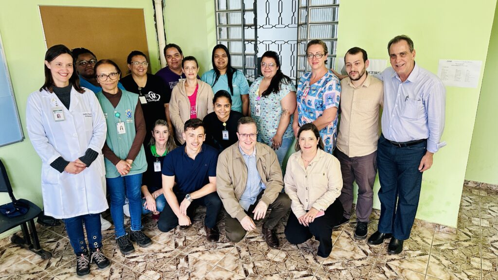 Na foto, há um grupo de 15 pessoas, sendo 12 mulheres e 3 homens. Eles estão em um ambiente interno, possivelmente uma clínica ou hospital, com paredes verdes e um chão de azulejos marrons e beges. No lado esquerdo da foto, uma mulher com jaleco branco está em destaque. Abaixo dela, há uma cadeira preta. Ao lado dela, uma mulher com colete verde, e atrás delas, outras mulheres com diferentes expressões e vestimentas. No centro da foto, um grupo de pessoas está mais agrupado. Um homem de jaqueta bege e óculos está agachado. Ao seu lado, outro homem de camisa clara e óculos está em pé, com o braço apoiado no ombro do homem ao seu lado. Há também mulheres com roupas coloridas e estampadas. No lado direito da foto, um homem de camisa azul clara e calça jeans escura está em pé, sorrindo. Ao seu lado, há uma mulher de blusa bege. Há também um aviso na parede com letras pretas. A iluminação da foto é boa, destacando os rostos e as vestimentas das pessoas. A imagem transmite uma atmosfera de equipe e colaboração.