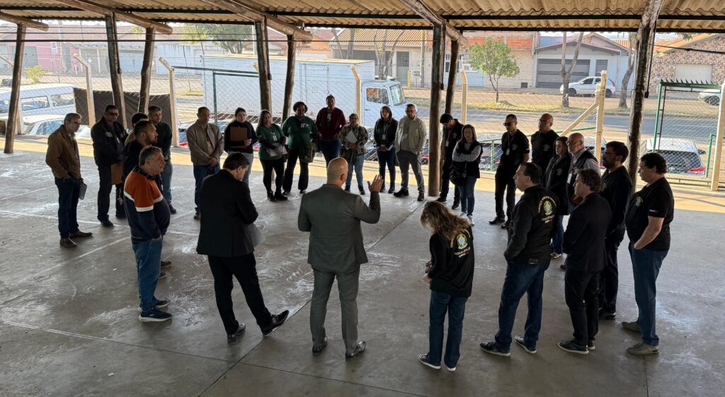 imagem mostra 30 pessoas reunidas em um circulo em uma quadra esportiva ou salão de festas. As pessoas prestam atenção na fala de uma homem, branco, vestido com roupas sociais e gesticula as mãos