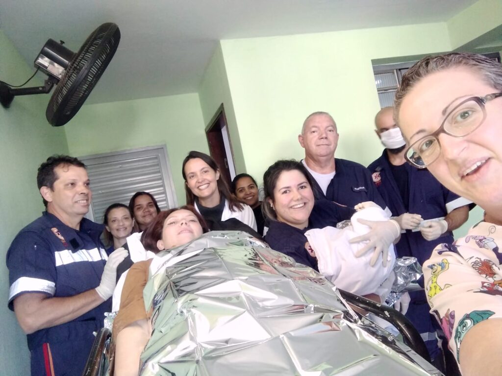 A imagem mostra um grupo de pessoas em um ambiente interno, possivelmente uma sala de emergência ou unidade médica. A maioria das pessoas parece ser pessoal médico, vestindo uniformes escuros.No centro da imagem, uma mulher está deitada em uma maca, coberta com um cobertor térmico prateado. Uma das pessoas, vestindo luvas, segura um recém-nascido enrolado em um cobertor branco.A atmosfera geral parece ser de sucesso e celebração, com muitos dos indivíduos sorrindo para a câmera. Um ventilador de teto é visível no canto superior esquerdo da imagem.