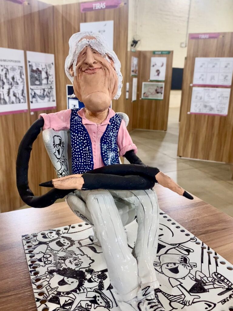 Escultura em cerâmica representando um homem idoso sentado, com cabelos brancos, colete azul de bolinhas sobre camisa rosa e calça cinza clara. A escultura está posicionada sobre uma mesa com desenhos em preto e branco, em um espaço de exposição.