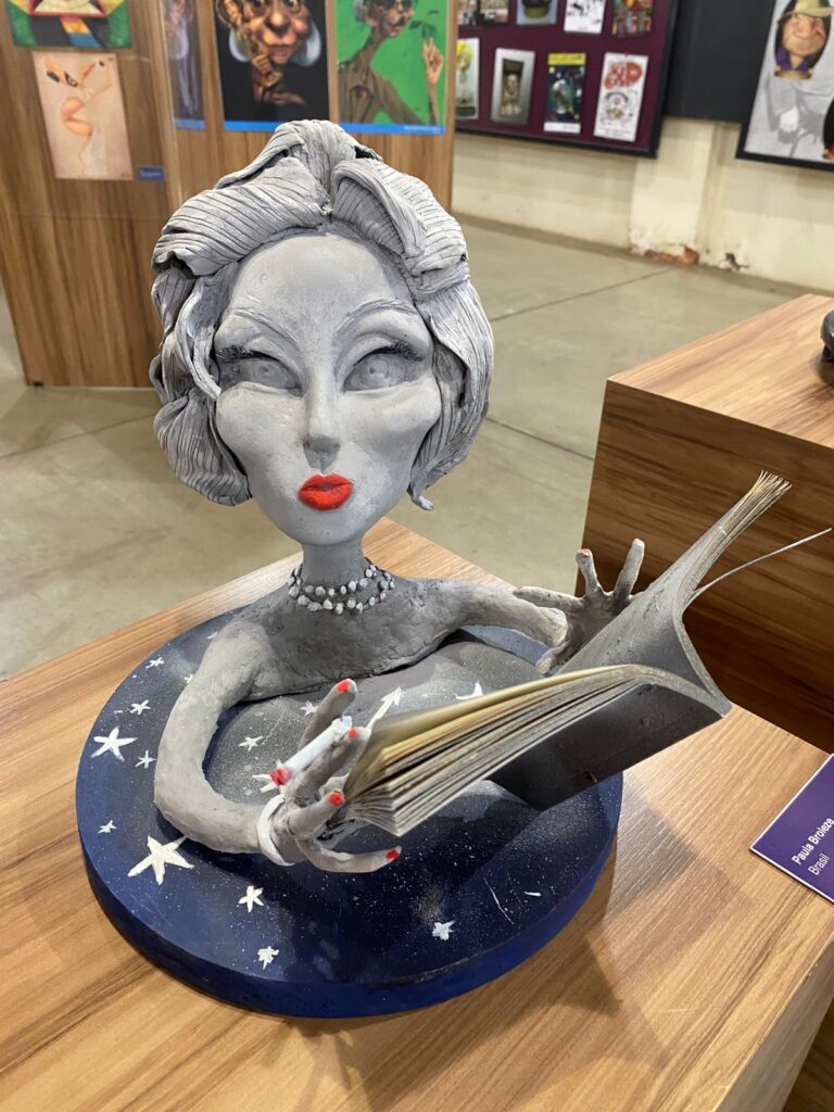 Escultura de Paula Broleze: busto feminino estilizado, em tom cinza, com livro aberto nas mãos.
