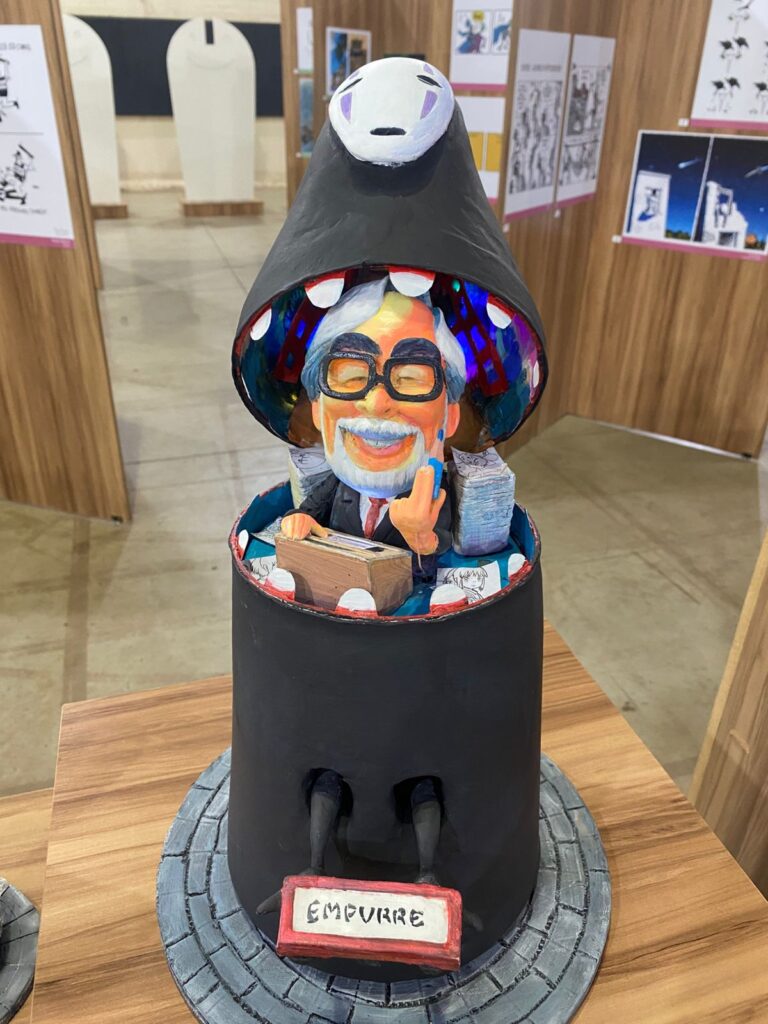 Escultura de Rui Miranda: figura caricatural saindo de dentro de um objeto cilíndrico, com a palavra “empurre”.