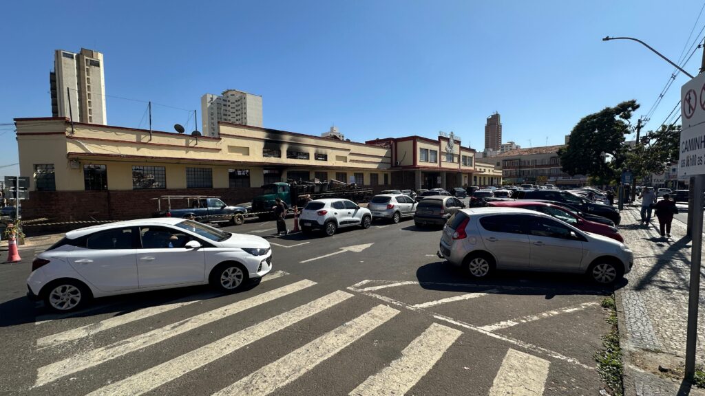 A imagem mostra uma movimentada cena urbana com carros estacionados e outros em movimento pela rua. Em destaque, há um edifício com áreas escurecidas que indicam danos causados por fogo ou fumaça, sugerindo um incêndio anterior. Ao fundo, edifícios altos completam o cenário típico de uma cidade grande.