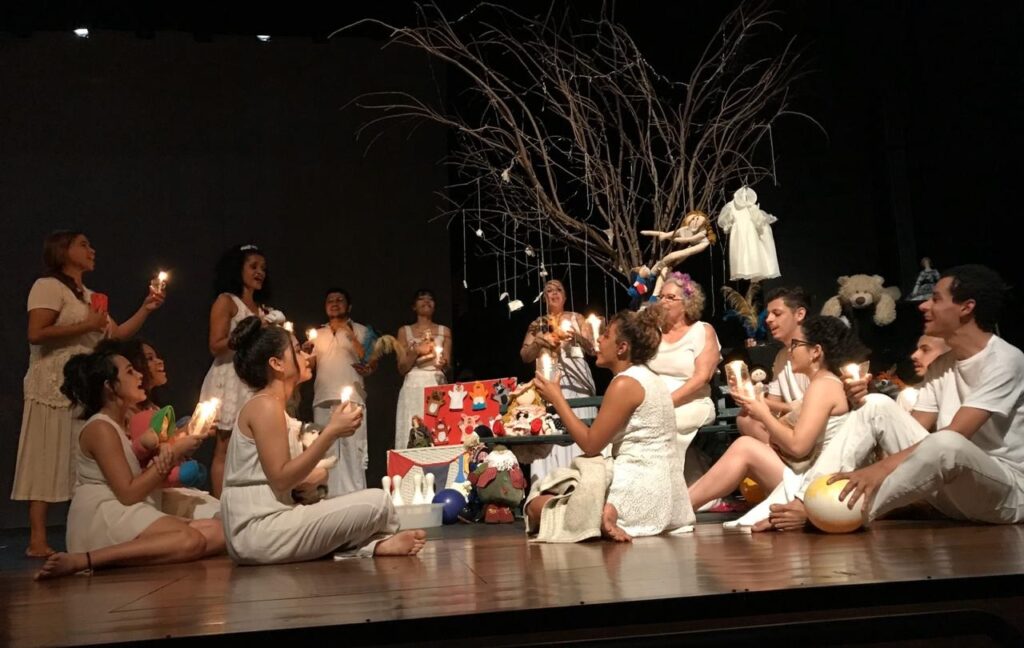 Cena de espetáculo teatral com elenco vestido de branco, segurando velas acesas, reunido em semicírculo diante de cenário com árvore seca decorada com bonecos.
