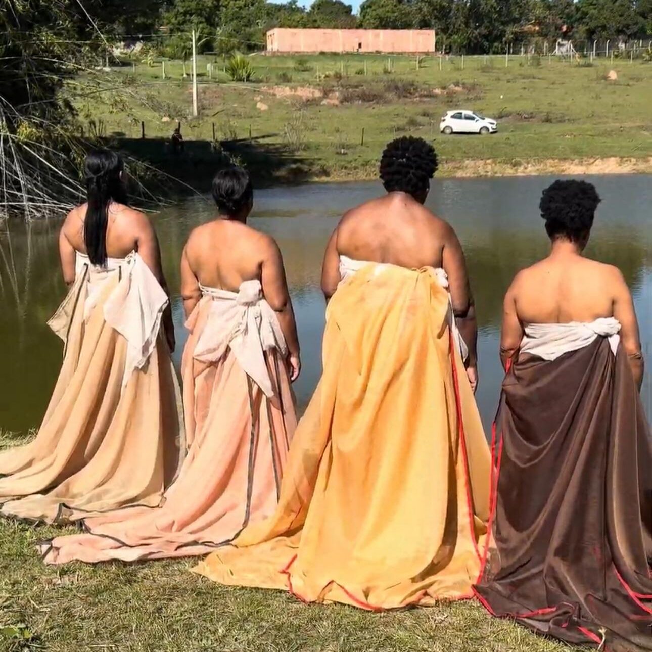 As mulheres estão em pé na grama, à beira de um lago, usando vestidos longos e fluidos em tons terrosos (bege, laranja, marrom). As costas estão nuas, destacando a conexão com a natureza. Ao fundo, uma paisagem rural com árvores, uma casa simples de alvenaria e um carro branco estacionado. A cena transmite ancestralidade, força e contemplação.