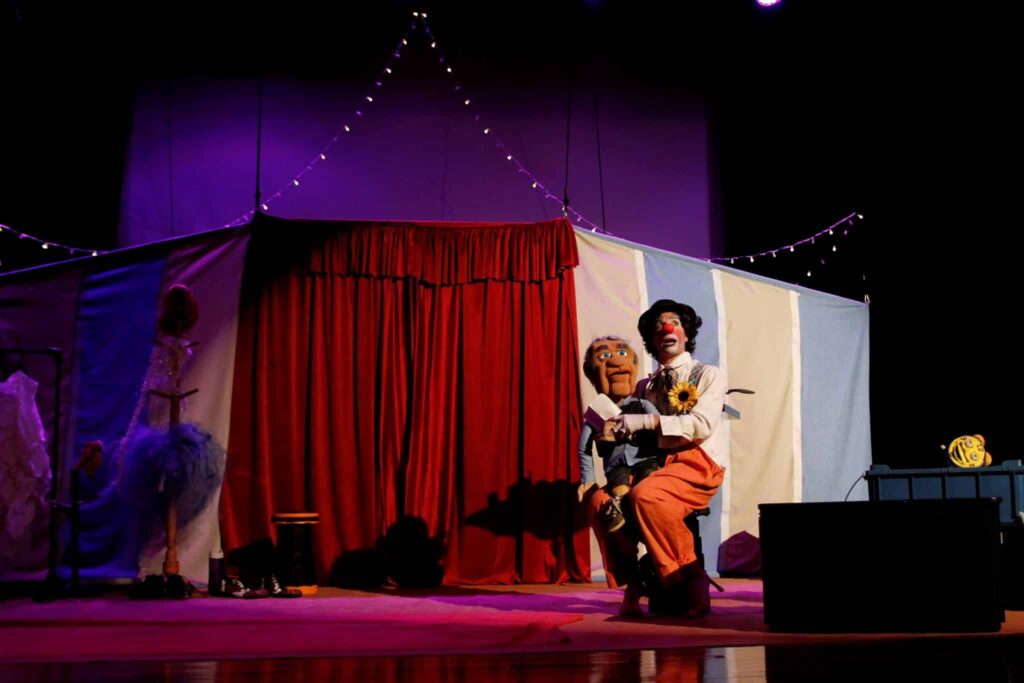 No palco de cortina vermelha e cenário que remete a uma lona de circo, o palhaço Picuinha aparece sentado com uma marionete no colo. Ele usa calça larga laranja, camisa branca com suspensórios coloridos, gravata com flor amarela e chapéu preto. O nariz vermelho e a maquiagem típica completam sua caracterização.