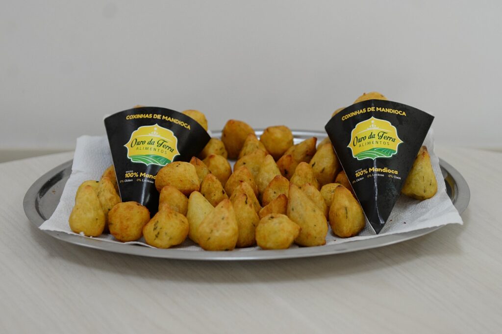 A imagem mostra uma travessa de metal cheia de coxinhas de mandioca douradas e apetitosas. As coxinhas estão dispostas em várias camadas e algumas parecem ter temperos verdes picados misturados na massa. No centro da travessa, há dois cones de papel preto com o logotipo "Ouro da Terra Alimentos". Os cones têm informações escritas em letras brancas, incluindo "COXINHAS DE MANDIOCA" na parte superior e "100% Mandioca", "0% Glúten", "0% Lactose" e "0% Ovos" na parte inferior. As informações nos cones sugerem que as coxinhas são feitas principalmente de mandioca e são livres de glúten, lactose e ovos. A travessa está colocada sobre uma superfície de madeira clara, e ao fundo, uma parede branca. A iluminação é suave, destacando a textura e a cor das coxinha