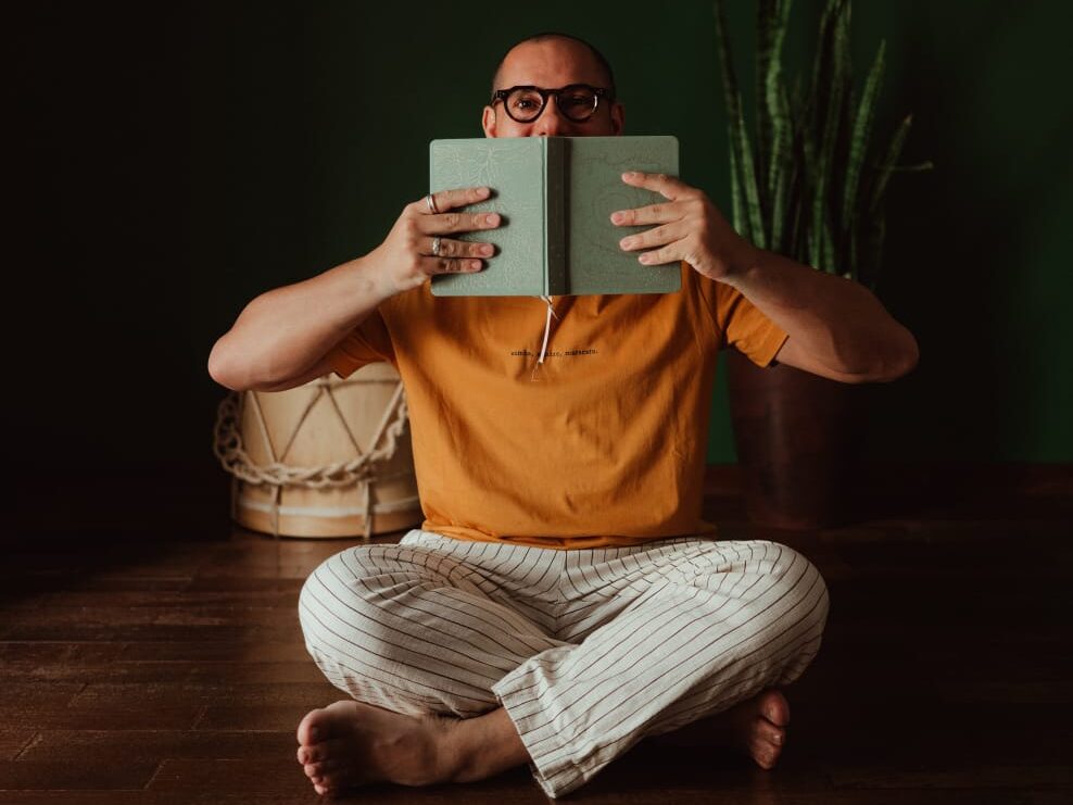 Homem careca, de óculos, está sentado no chão de pernas cruzadas, usando camiseta amarela e calça clara listrada. Ele segura um livro verde aberto na frente do rosto, cobrindo parte do sorriso. Atrás dele, há uma parede verde-escura, um vaso com planta e um tambor de madeira.