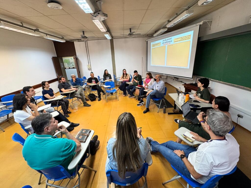 A imagem apresenta uma sala de aula organizada em formato circular, com cerca de 18 participantes sentados em cadeiras azuis com pranchetas acopladas, sugerindo um ambiente voltado à troca de ideias e à construção coletiva. O piso amarelo e as paredes brancas criam um espaço simples e funcional, enquanto um quadro verde e uma tela de projeção compõem os recursos visuais da sala.