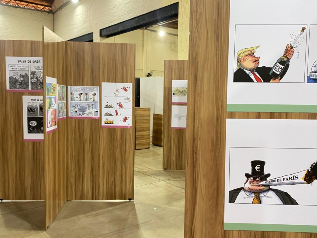 Vista de painéis de madeira clara organizados em um grande salão expositivo. Neles estão colados cartuns e tiras de humor com temáticas políticas e sociais, incluindo uma caricatura de Donald Trump e outro cartum sobre o Acordo de Paris.