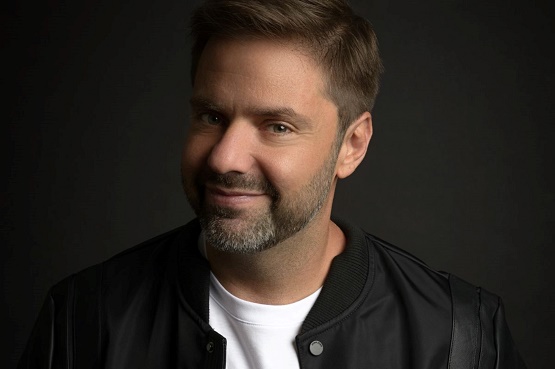 Fotografia de um homem sorridente de barba baixa e cabelo castanho claro, penteado para o lado. Ele veste uma jaqueta preta sobre camiseta branca, posando contra um fundo escuro.