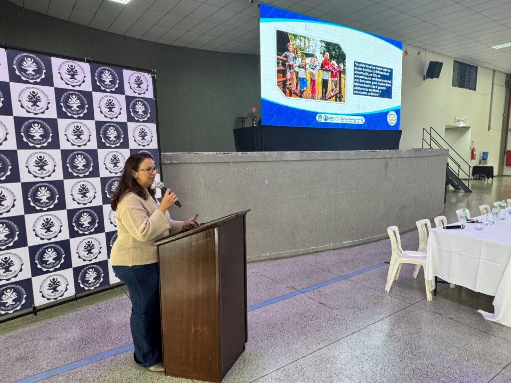 A mulher está em pé atrás de um púlpito de madeira, segurando um microfone em sua mão direita e uma caneta na esquerda. Ela está usando um suéter bege e jeans azuis. Atrás dela, há um banner com um padrão xadrez e o logotipo de uma organização. Na frente dela, há uma grande tela exibindo uma foto de crianças e texto. A sala parece ser um local de eventos, com mesas e cadeiras à direita.