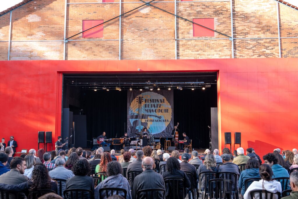 Foto mostra o palco do Teatro do Engenho, com fachada vermelha e parede de tijolos aparentes ao fundo. No centro, cinco músicos se apresentam no Festival de Jazz Manouche de Piracicaba, tocando guitarra, contrabaixo acústico, violão e bateria. A plateia, formada por dezenas de pessoas sentadas em cadeiras pretas, assiste atentamente.
