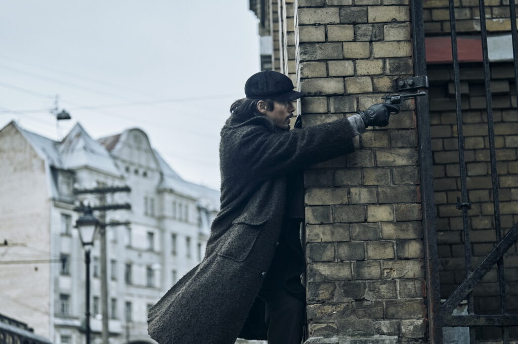 Homem usando casaco pesado e boné encosta-se a uma parede de tijolos e aponta uma arma para fora, em um cenário urbano de época.
