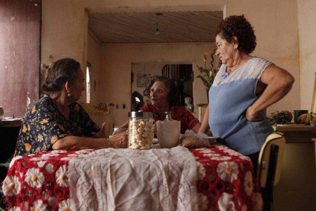 Pra Cego Ver: Três mulheres mais velhas conversam em volta de uma mesa coberta com toalha de crochê branca e vermelha. No centro da mesa há potes de vidro com biscoitos e alimentos. A personagem principal, ao centro, gesticula com expressão intensa, enquanto as outras duas escutam, em um ambiente simples de casa antiga.