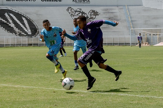 A imagem mostra uma partida de futebol entre jovens jogadores em um campo gramado sob um céu ensolarado. Dois jogadores estão em primeiro plano, em plena ação. O jogador à esquerda, vestindo um uniforme azul claro com um padrão intrincado, corre com a bola nos pés, com uma expressão de concentração no rosto. O jogador à direita, com um uniforme roxo escuro, está em movimento, com o braço estendido para alcançar a bola. Ambos os jogadores usam chuteiras e meias altas. Ao fundo, outros jogadores podem ser vistos em campo, e as arquibancadas de um estádio, parcialmente visíveis, sugerem que se trata de um evento esportivo. O campo está bem cuidado, com linhas brancas marcando os limites. A iluminação forte indica que é dia, e a sombra projetada pelos jogadores reforça essa percepção. A cena captura a energia e a intensidade de um jogo de futebol juvenil.
