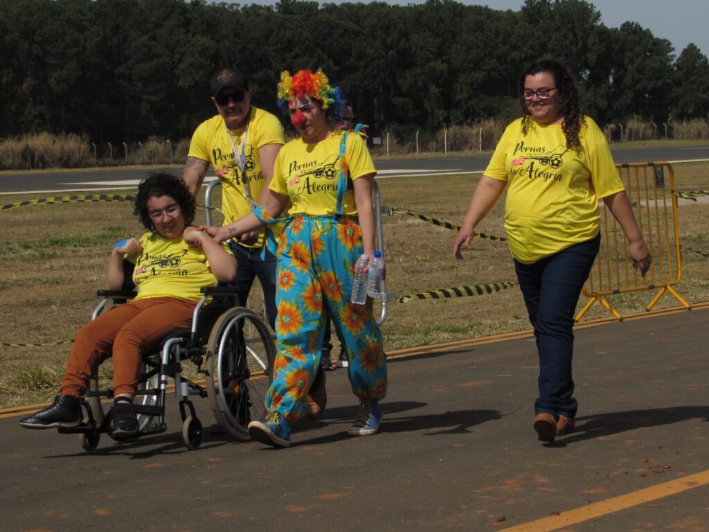 Grupo caminha ao ar livre; uma mulher empurra jovem em cadeira de rodas, acompanhada por pessoas vestindo camisetas amarelas, incluindo uma palhaça de peruca colorida