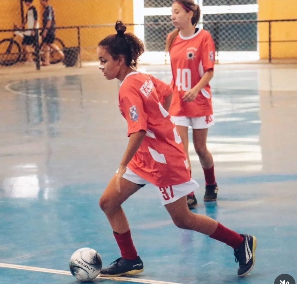 Na imagem, duas jovens jogadoras de futsal estão em ação em uma quadra com piso azul. A jogadora em primeiro plano, com o número 37 em sua camisa vermelha com detalhes brancos, está driblando a bola com concentração. Ela tem cabelos escuros presos em um coque e usa meias vermelhas compridas e chuteiras pretas com detalhes em amarelo.Ao fundo, outra jogadora, de número 40 em sua camisa semelhante, observa a jogada. Ela tem cabelos claros e também veste meias vermelhas e chuteiras. A cena sugere um momento dinâmico de uma partida de futsal, com o ambiente da quadra e outros elementos desfocados ao fundo, como bicicletas e uma grade.