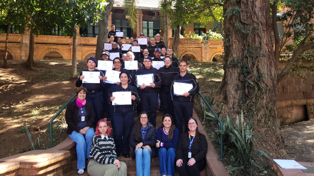 A imagem mostra um grupo de pessoas sentadas e em pé em degraus de tijolos, com uma grande árvore à direita e um edifício de tijolos ao fundo.A maioria das pessoas está vestindo uniformes escuros, possivelmente uniformes de trabalho ou de uma organização. Vários indivíduos estão segurando certificados ou documentos. O grupo parece estar participando de uma cerimônia de formatura ou de um evento de reconhecimento.As pessoas estão dispostas em várias fileiras nos degraus, com algumas em pé atrás das que estão sentadas. A iluminação sugere que a foto foi tirada durante o dia, com luz solar entrando por entre as árvores. A atmosfera parece ser de celebração e conquista.