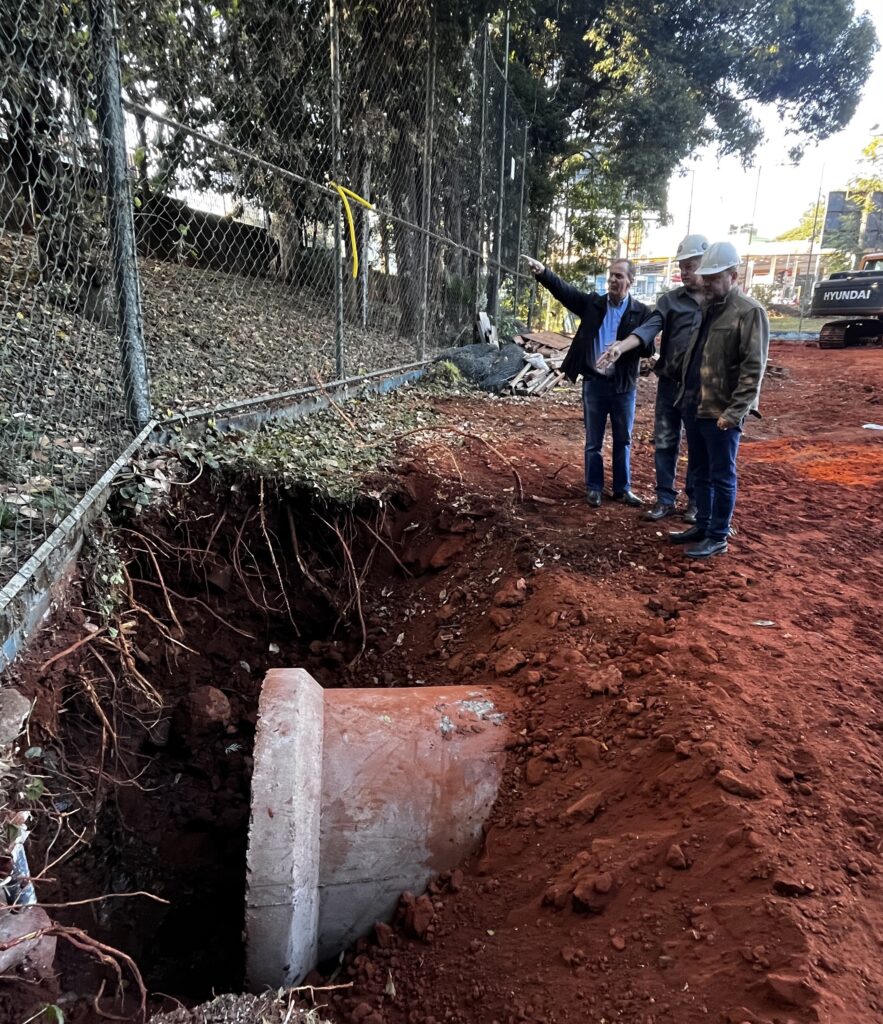 A imagem mostra três homens vestidos com roupas casuais e capacetes de segurança inspecionando uma obra. Eles estão em pé em um terreno com terra vermelha e há uma grande escavação no chão, revelando um tubo de concreto laranja. O homem mais à esquerda, vestindo uma camisa azul e uma jaqueta escura, está apontando com o dedo indicador para algo fora do quadro. Os outros dois homens, vestidos com jaquetas, estão de pé ao lado dele, observando na mesma direção. Ao fundo, pode-se ver uma cerca de arame, árvores e um trator com a marca "HYUNDAI". A cena sugere que os homens estão supervisionando o progresso da construção, possivelmente discutindo o trabalho que está sendo feito na tubulação exposta.