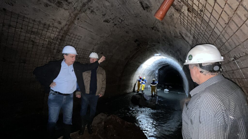 A imagem mostra um grupo de pessoas em um túnel de concreto. No primeiro plano, à esquerda, um homem com capacete branco e blazer preto sobre uma camisa azul clara está de pé, com a mão no quadril e a outra estendida, como se estivesse apontando para algo no túnel. Ele veste calças jeans e botas de borracha. À direita, no primeiro plano, outro homem com capacete branco e uma camisa xadrez está de costas, olhando para o túnel. O capacete dele tem um adesivo verde com um símbolo. No fundo do túnel, há outras pessoas vestindo equipamentos de segurança, incluindo capacetes e macacões amarelos e azuis. Elas parecem estar trabalhando ou inspecionando o túnel. O túnel é feito de concreto e tem uma estrutura de metal aparente em algumas partes do teto e das paredes. Há água corrente no chão do túnel, e uma mangueira laranja está pendurada no teto, com água pingando dela. A iluminação no túnel vem de algumas luzes no teto e da entrada mais adiante. A atmosfera geral sugere uma inspeção ou trabalho em uma infraestrutura subterrânea.