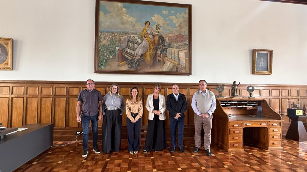 A imagem retrata seis pessoas em pé em uma sala com painéis de madeira nas paredes e piso de parquet, sugerindo um ambiente formal e histórico. Ao fundo, destaca-se uma grande pintura de uma mulher sentada em uma máquina, possivelmente ligada à indústria têxtil, com uma paisagem ao fundo. Flanqueando a pintura, há dois retratos emoldurados — um à esquerda e outro acima de uma escrivaninha de madeira com gavetas e tampo retrátil à direita. Sobre essa escrivaninha, observam-se itens decorativos e uma pequena estátua. À esquerda, uma mesa moderna com papéis reforça o uso atual do espaço, que parece ser um escritório ou sala de reuniões com valor simbólico e institucional.
