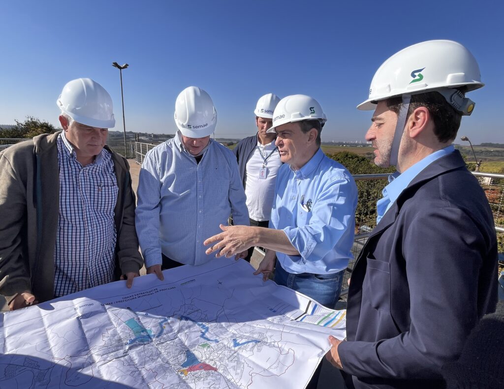 Um grupo de cinco homens, todos usando capacetes brancos, está reunido em torno de um grande mapa desdobrado. Eles parecem estar em uma área externa, com um céu azul claro e algumas árvores e edifícios ao fundo. Os homens estão vestidos de forma casual e profissional. Quatro deles usam camisas de manga comprida, sendo duas azuis claras e uma xadrez. O homem mais à direita usa um blazer escuro sobre uma camisa azul clara. O homem mais à esquerda usa uma jaqueta marrom sobre uma camisa xadrez. O homem no centro, usando uma camisa azul clara, está gesticulando com a mão direita sobre o mapa, explicando algo aos outros. Os outros homens estão olhando atentamente para o mapa, alguns inclinados para frente para ver melhor. O mapa é grande e detalhado, com várias linhas, cores e símbolos. Na parte inferior do mapa, é possível ler "OBRAS ESTRATÉGICAS" e "SEMAE / PREFEITURA MUNICIPAL DE PIRACICABA". Os capacetes de alguns homens têm o logotipo "SEMAE" em branco e verde, indicando que eles podem estar envolvidos com essa organização. A cena sugere uma reunião de planejamento ou inspeção de um projeto de infraestrutura, possivelmente relacionado a obras estratégicas da prefeitura municipal de Piracicaba.