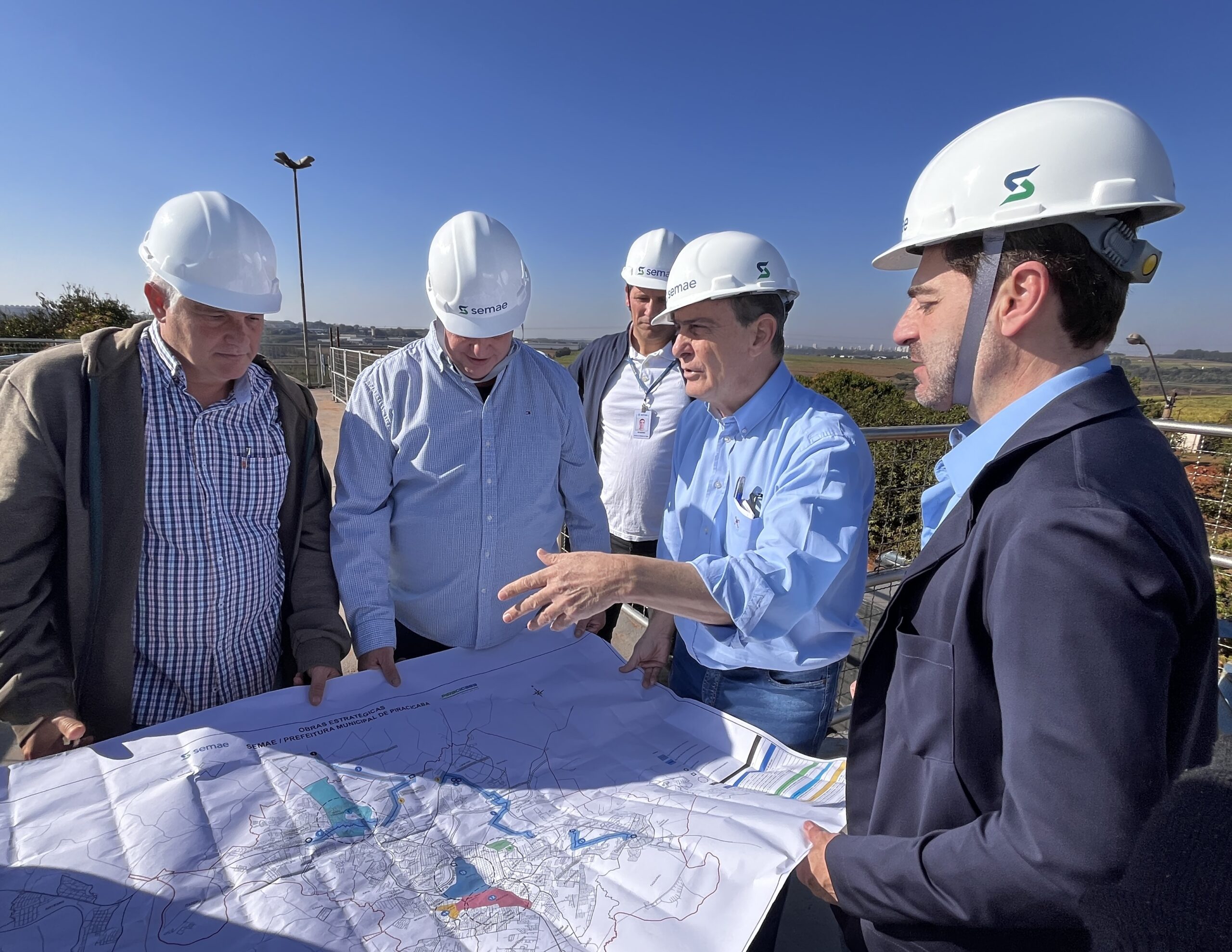 Um grupo de cinco homens, todos usando capacetes brancos, está reunido em torno de um grande mapa desdobrado. Eles parecem estar em uma área externa, com um céu azul claro e algumas árvores e edifícios ao fundo. Os homens estão vestidos de forma casual e profissional. Quatro deles usam camisas de manga comprida, sendo duas azuis claras e uma xadrez. O homem mais à direita usa um blazer escuro sobre uma camisa azul clara. O homem mais à esquerda usa uma jaqueta marrom sobre uma camisa xadrez. O homem no centro, usando uma camisa azul clara, está gesticulando com a mão direita sobre o mapa, explicando algo aos outros. Os outros homens estão olhando atentamente para o mapa, alguns inclinados para frente para ver melhor. O mapa é grande e detalhado, com várias linhas, cores e símbolos. Na parte inferior do mapa, é possível ler "OBRAS ESTRATÉGICAS" e "SEMAE / PREFEITURA MUNICIPAL DE PIRACICABA". Os capacetes de alguns homens têm o logotipo "SEMAE" em branco e verde, indicando que eles podem estar envolvidos com essa organização. A cena sugere uma reunião de planejamento ou inspeção de um projeto de infraestrutura, possivelmente relacionado a obras estratégicas da prefeitura municipal de Piracicaba.