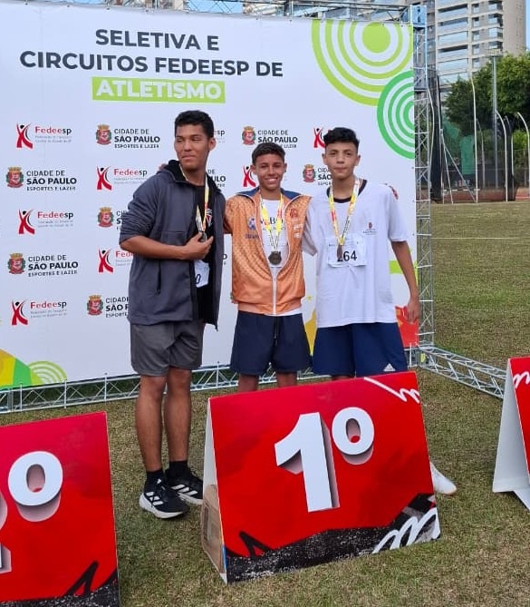 A imagem mostra três jovens atletas em um pódio, comemorando suas vitórias em um evento de atletismo. Eles estão vestindo medalhas e sorrindo para a câmera. Atrás deles, há um grande banner com o título "Seletiva e Circuitos Fedeesp de Atletismo" e os logotipos da Fedeesp e da Cidade de São Paulo. Os pódios para os primeiro, segundo e terceiro lugares estão visíveis na frente dos atletas. O jovem no centro, vestindo uma jaqueta laranja, parece ter conquistado o primeiro lugar, pois está posicionado no centro do pódio e ostenta uma medalha maior. Os outros dois jovens estão ao seu lado, também com medalhas. O evento parece ter sido realizado ao ar livre, em um campo gramado.