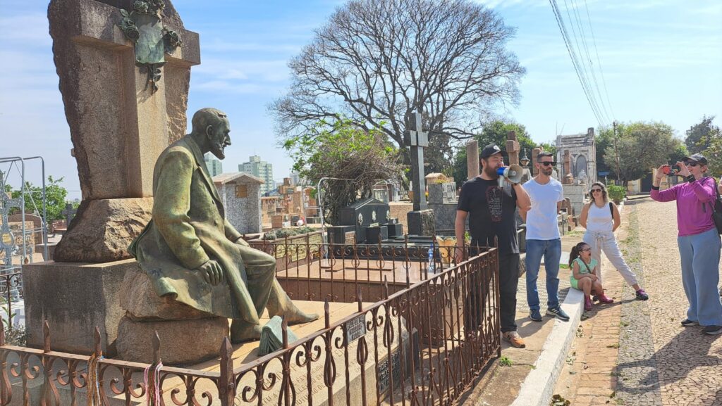 A imagem mostra um grupo de pessoas em um cemitério, em um dia ensolarado com céu azul e algumas nuvens.No primeiro plano, à esquerda, há um grande monumento de pedra em formato de cruz, com uma estátua de bronze de um homem sentado, vestindo terno e gravata. A estátua está virada para a direita e parece estar olhando para o horizonte. Há uma grade de metal enferrujada em frente ao monumento.Um grupo de cinco pessoas está em pé em uma calçada de paralelepípedos, atrás da grade. O homem mais à esquerda está usando um boné preto e uma camiseta preta com uma estampa, e está segurando um megafone, como se estivesse falando com o grupo. Ao lado dele, um homem com óculos escuros e camiseta branca está olhando para ele. Mais atrás, uma mulher com óculos de sol e uma blusa branca está olhando para o grupo. Uma menina pequena, vestida com uma roupa verde clara, está sentada em um meio-fio, bocejando. No lado direito da imagem, uma mulher com um boné, óculos escuros e uma blusa rosa está tirando uma foto com o celular.Ao fundo, o cemitério se estende, com várias lápides, cruzes e outros monumentos funerários. Uma grande árvore com galhos secos domina o centro do fundo. Ao longe, prédios altos podem ser vistos. O sol brilha intensamente, criando sombras nítidas no chão.