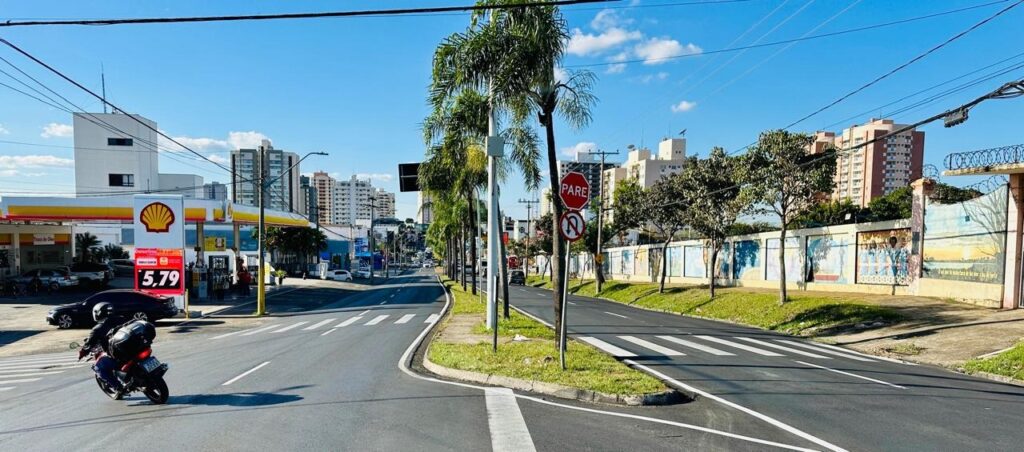 A imagem mostra uma rua com vários prédios altos ao fundo, sob um céu azul claro com algumas nuvens. Há uma estação de serviço Shell à esquerda, com um preço de gasolina exibido em um painel. Uma motocicleta com um piloto está se movendo na rua. Há uma faixa de pedestres na estrada, e as árvores de palma alinham o lado direito da rua. Uma placa de "PARE" é visível em um poste de luz. Uma parede com murais coloridos está ao longo do lado direito da rua.