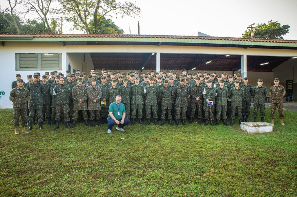 A imagem mostra um grupo de jovens em uniformes militares, em pé em um gramado em frente a um edifício. Um homem de camiseta verde está agachado na frente do grupo. O grupo parece estar posando para uma foto.