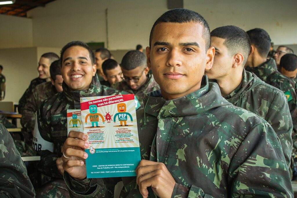 A imagem mostra um grupo de jovens em uniformes militares, possivelmente soldados ou cadetes, alinhados. O foco está nos dois homens em primeiro plano. O homem à esquerda está sorrindo e olhando para o espectador, enquanto o homem à direita está olhando diretamente para a câmera com uma expressão neutra. O homem à direita segura um folheto colorido que possui texto e ilustrações em estilo de cartoon. O título do folheto é "Os nomes são parecidos, mas há diferenças". Abaixo, há duas figuras, uma rotulada como "PrEP" (em azul) e a outra como "PEP" (em laranja), com ilustrações de vírus entre elas. O texto discute a PrEP (Profilaxia Pré-Exposição) e a PEP (Profilaxia Pós-Exposição) em relação ao HIV. Na parte inferior do folheto, há informações de contato do CEDIC - Centro de Doenças Infecto Contagiosas. Os outros indivíduos ao fundo também estão em uniforme e parecem fazer parte do mesmo grupo, com alguns olhando para frente e outros olhando para baixo. O ambiente parece ser uma instalação de treinamento ou educacional, com mesas visíveis ao fundo. A impressão geral é a de uma sessão de conscientização ou educativa sendo realizada em um contexto militar.