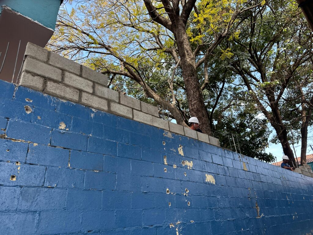 Nesta imagem, vemos uma parede parcialmente construída com blocos de concreto sobre uma base de tijolos pintados de azul, cuja pintura apresenta desgaste e descascamento, sugerindo exposição prolongada ao tempo. Atrás da parede, um trabalhador com capacete branco de segurança está parcialmente visível, indicando que a obra está em andamento. Ao fundo, há árvores altas com folhas verdes e amareladas, além de um edifício de fachada rosada parcialmente visível à esquerda.