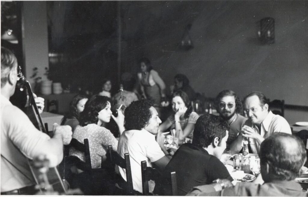 Registro em preto e branco de um encontro em restaurante. Jaguar está sentado entre Ziraldo, Fortuna, Zélio e outros cartunistas, discutindo animadamente sobre o futuro Salão Internacional de Humor de Piracicaba. Ao fundo, músicos animam a reunião.