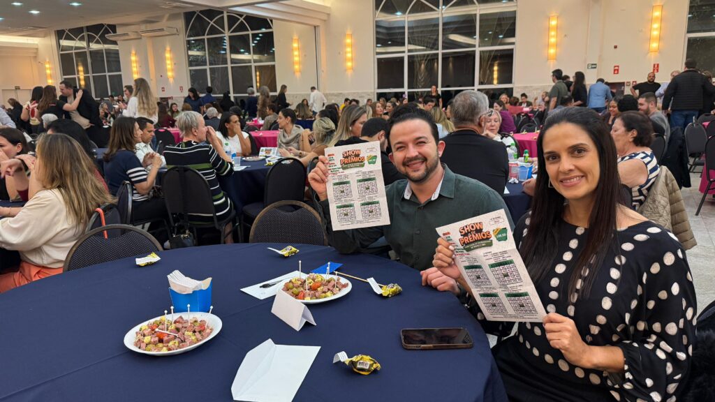 A imagem mostra um evento social em um salão com várias mesas redondas. Duas pessoas estão em primeiro plano, sentadas à mesa, segurando cartelas de bingo com o título "SHOW DE PRÊMIOS". O homem, à esquerda, veste uma camisa verde escura e sorri para a câmera. A mulher, à direita, usa um vestido preto com bolinhas brancas e também sorri. Na mesa à frente deles, há um prato com petiscos, guardanapos e um celular. O salão está cheio de pessoas, a maioria sentada em mesas, conversando e participando do evento. Há iluminação artificial no teto e janelas grandes com vista para o exterior, embora a imagem externa não seja clara. O ambiente parece ser de confraternização e entretenimento.