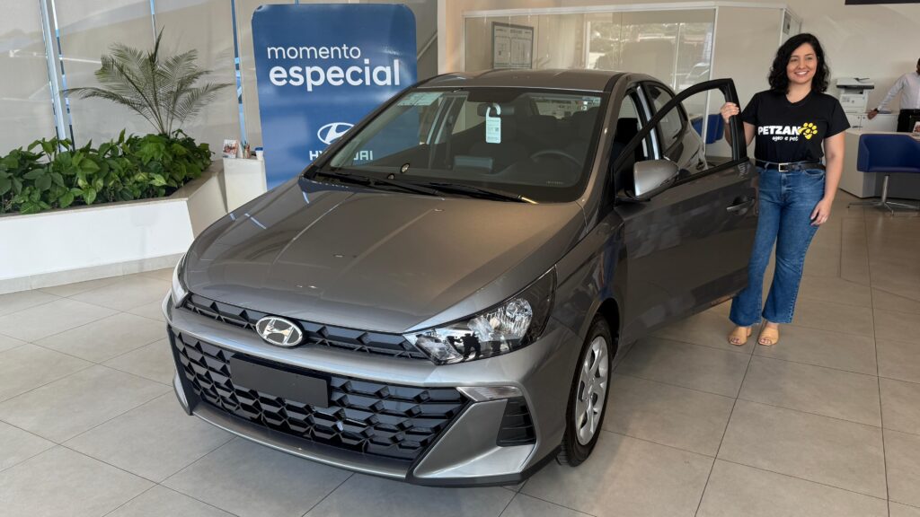 A imagem mostra uma mulher em pé ao lado de um carro cinza em uma concessionária. Ela está vestindo uma camiseta preta com o logotipo "PETZAN agro e pet" e jeans azul. A porta do motorista do carro está aberta e ela está apoiada nela. Atrás dela, há um banner azul com as palavras "momento especial" e o logotipo da Hyundai. O carro parece ser um modelo novo, com um design moderno e grade frontal proeminente. O interior da concessionária é iluminado e limpo, com plantas verdes e móveis de escritório visíveis ao fundo.