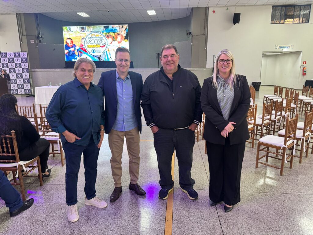 A imagem mostra quatro pessoas em pé em um evento. Da esquerda para a direita, um homem com cabelo loiro comprido e camisa azul escura, um homem com óculos e blazer azul escuro sobre uma camisa azul clara, um homem mais corpulento com jaqueta preta e jeans escuros, e uma mulher com óculos, blazer preto e uma blusa estampada. Todos sorriem para a câmera.