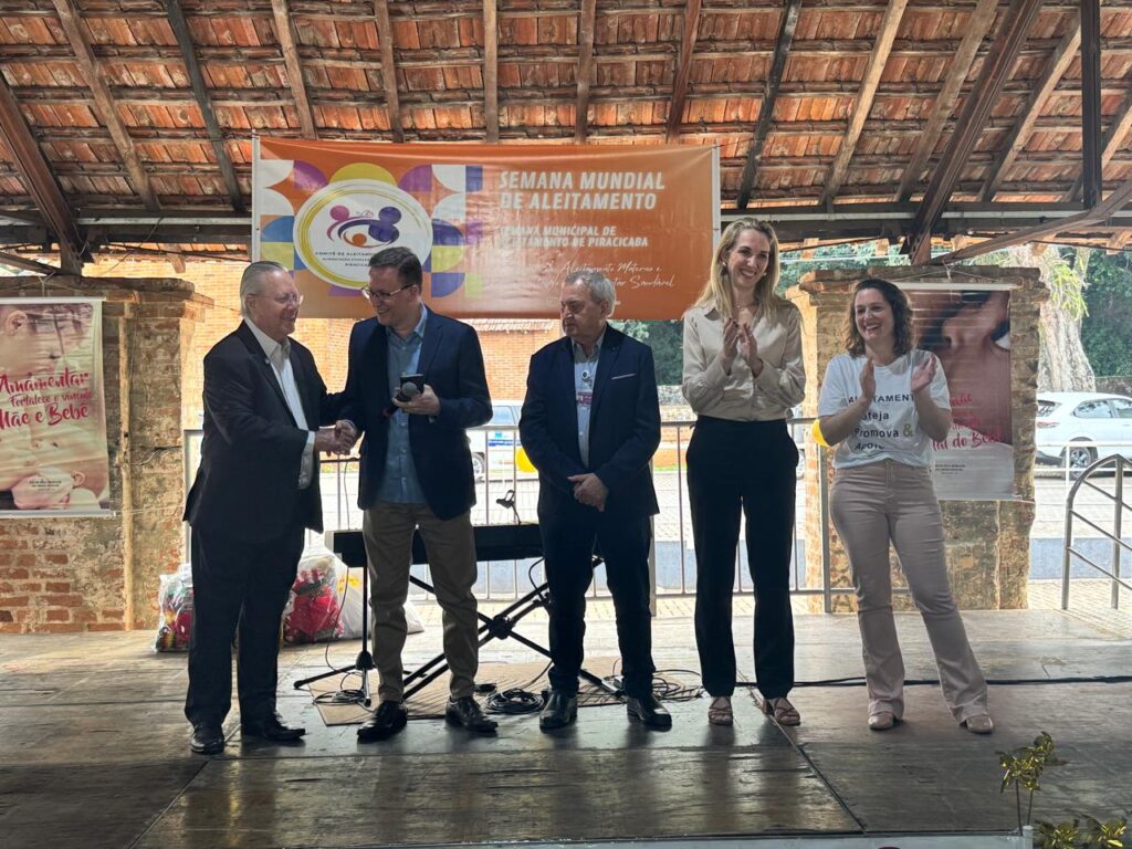 O evento retratado na imagem é a Semana Mundial de Aleitamento, com foco municipal em Piracicaba. A celebração, que visa fortalecer o vínculo entre mãe e bebê, conta com a presença de diversas personalidades, incluindo autoridades e profissionais de saúde. No palco, um homem mais velho, vestindo terno escuro, aperta a mão de um homem mais jovem, que usa terno azul e segura um microfone. Ambos sorriem, indicando um momento de reconhecimento ou parceria. Atrás deles, um banner laranja exibe o tema do evento e o logotipo do Comitê de Aleitamento. Duas mulheres, à direita, também aplaudem. Uma delas, com um vestido claro, segura um microfone e parece discursar, enquanto a outra, com uma camiseta branca, sorri e bate palmas. O ambiente parece ser ao ar livre, com uma estrutura de madeira no teto e árvores ao fundo. A imagem transmite uma atmosfera de celebração e compromisso com a promoção do aleitamento materno.
