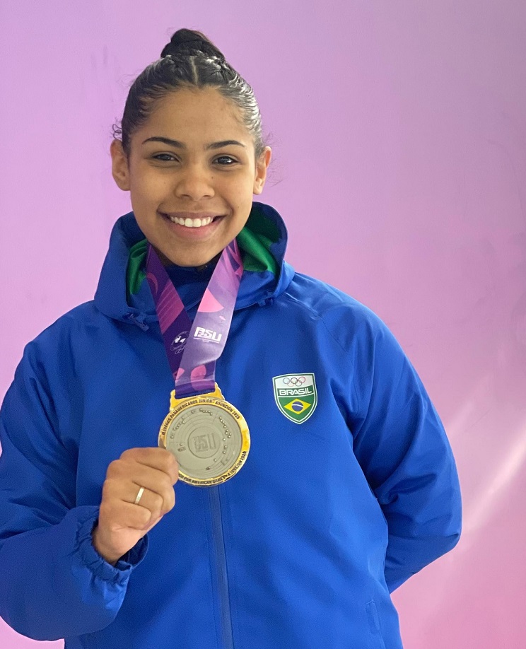A jovem sorri radiante para a câmera, exibindo uma medalha de prata pendurada em seu pescoço. Ela veste uma jaqueta azul com o emblema do Brasil no lado direito do peito. A medalha, com um cordão roxo e rosa, ostenta as inscrições "PSU" e "JOGOS PANAMERICANOS JUNIOR ASUNCIÓN 2029". O fundo da imagem é um tom suave de lilás.