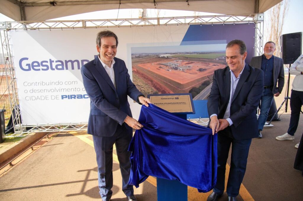 A imagem mostra um momento simbólico de inauguração, onde dois homens estão revelando uma placa coberta por um tecido azul. Ao fundo, há um banner com o logotipo da empresa Gestamp e a frase “Contribuindo com o desenvolvimento da CIDADE DE PIRACICABA”, reforçando o compromisso institucional com a região. Também é visível uma grande fotografia de um canteiro de obras, sugerindo que o evento marca o início de um novo empreendimento. A placa revelada traz os dizeres “Gestamp PEDRA FUNDAMENTAL”, indicando o lançamento oficial da construção.